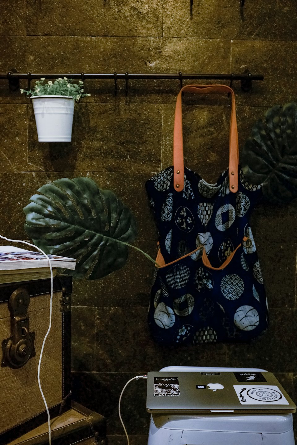 A tote bag hangs on a stone wall near a laptop on a stool.