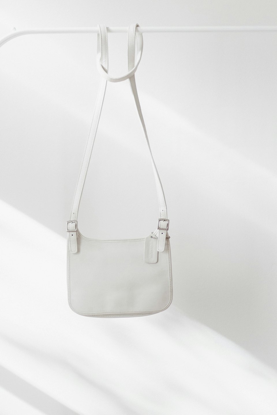 White leather crossbody bag with silver buckles hanging on a rail.